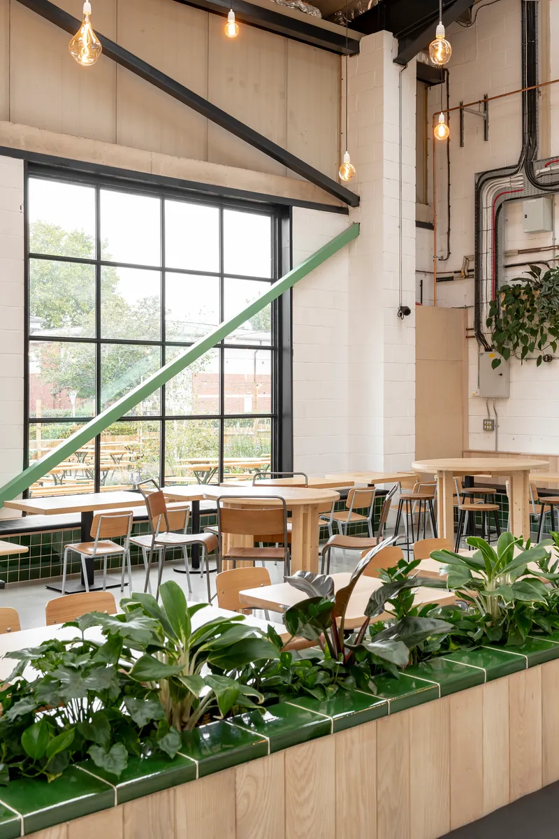 Double-height dining space with exposed concrete columns, large gridded windows and planted timber booth dividers