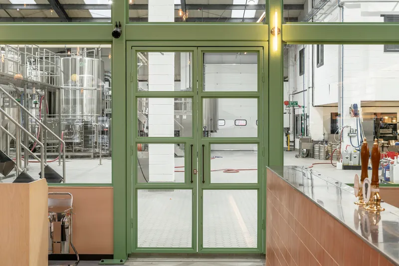Green framed glass partition separating dining area from brewery equipment and stainless steel tanks beyond
