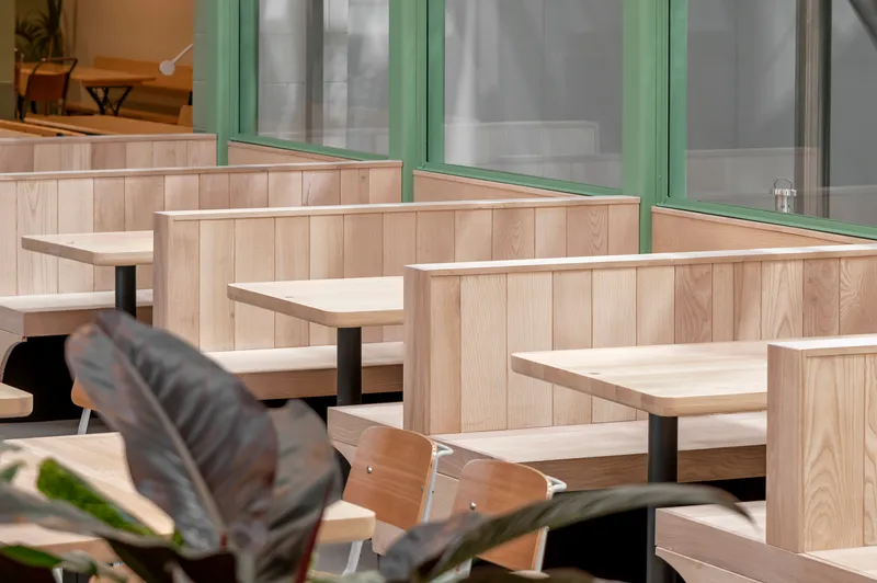Close-up of light timber booth seating with vertical wood paneling and fixed tabletops near plant leaves