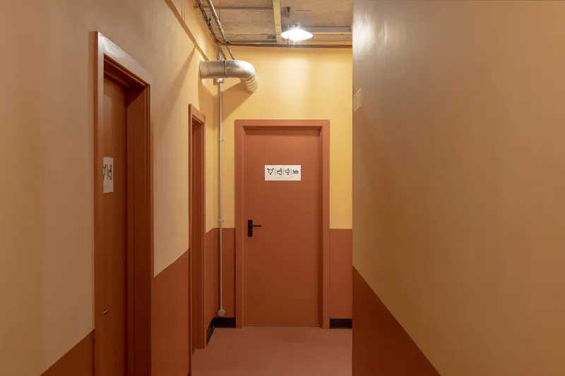 Corridor with terracotta and cream painted walls leading to a restroom door beneath exposed services