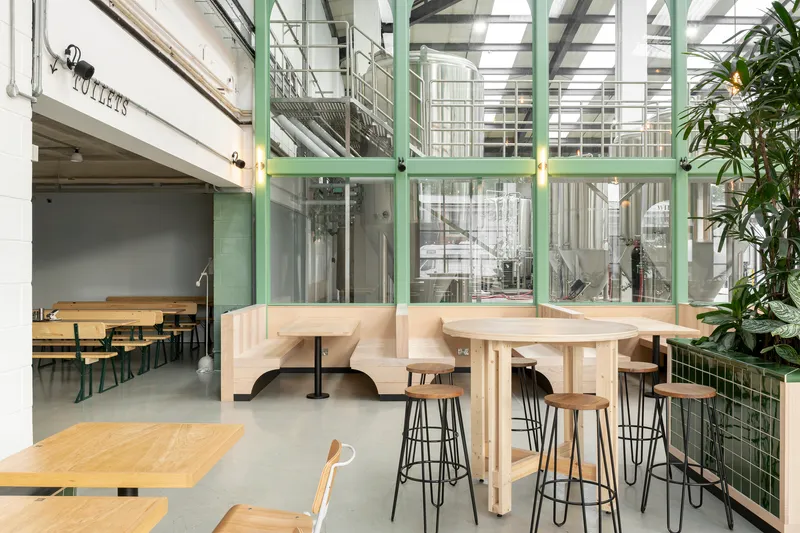 Interior dining space with pale timber booth seating and green-framed glass partitions revealing industrial brewery equipment beyond