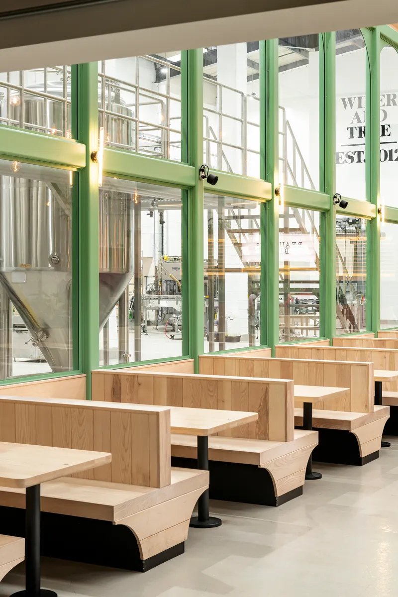 Row of pale timber booth seats beside green-framed glazing with views into the brewery production area