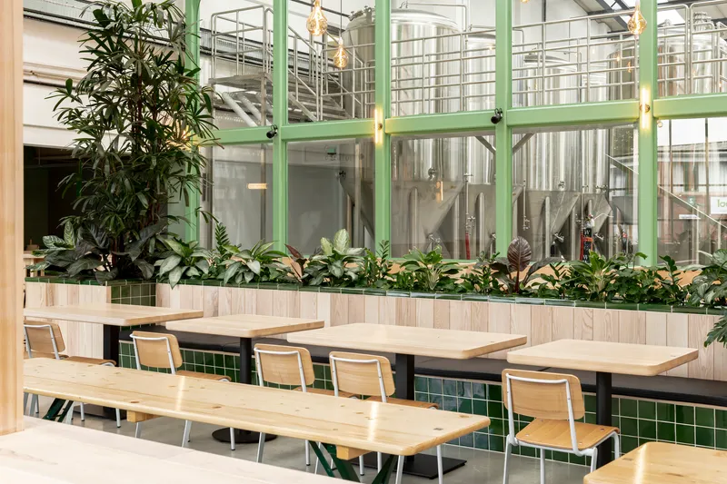 Long timber tables and benches beside a raised planter bed with steel brewing vessels beyond