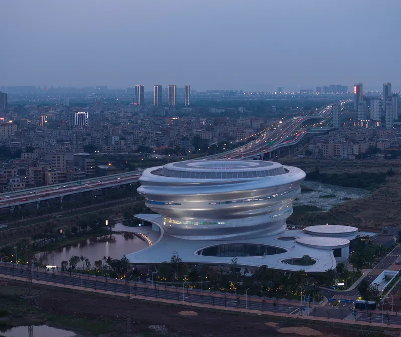 Aerial view of the spiraling ribbed volume beside a highway at dusk with city lights illuminating the background