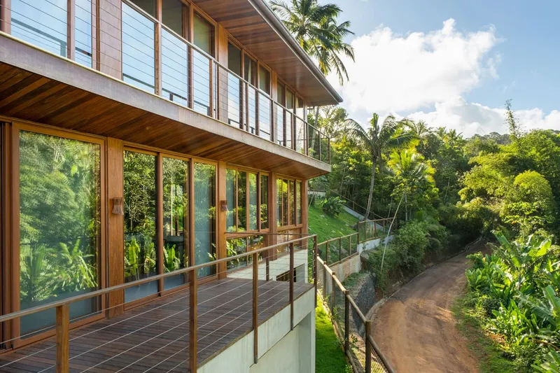 Two-story timber facade with glass balcony railing overlooking forested slope and dirt road