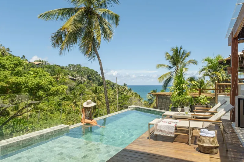 Infinity pool deck with timber loungers overlooking palm trees and the ocean beyond under clear skies