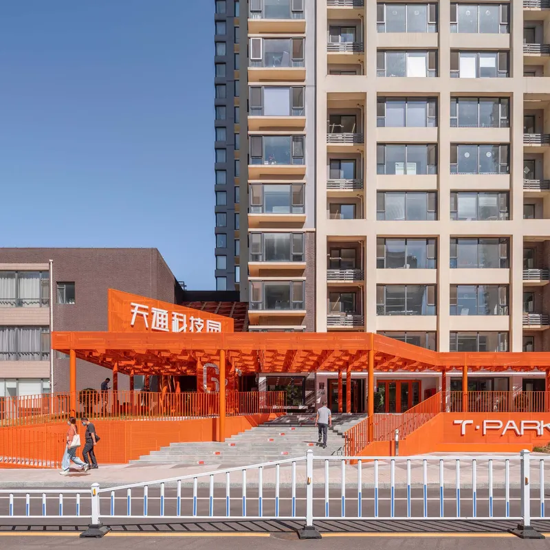 Orange lattice canopy over plaza entrance with pedestrians passing street barrier in daylight