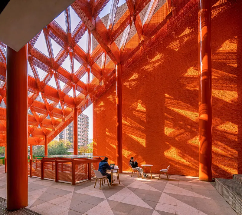 Interior view beneath orange perforated metal screen casting shadow patterns on tiled floor with seated figures