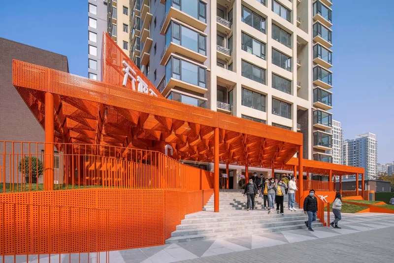 Perforated orange steel canopy with geometric underside sheltering visitors near residential towers