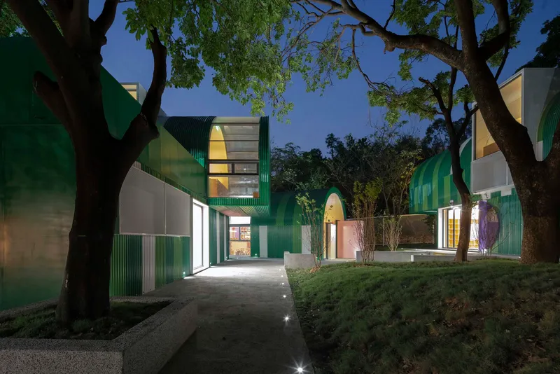 Illuminated pathway between green paneled volumes under mature trees at twilight