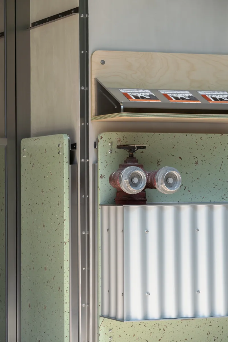 Close-up of exposed water valve and pipes mounted on pale green terrazzo wall beneath plywood shelf