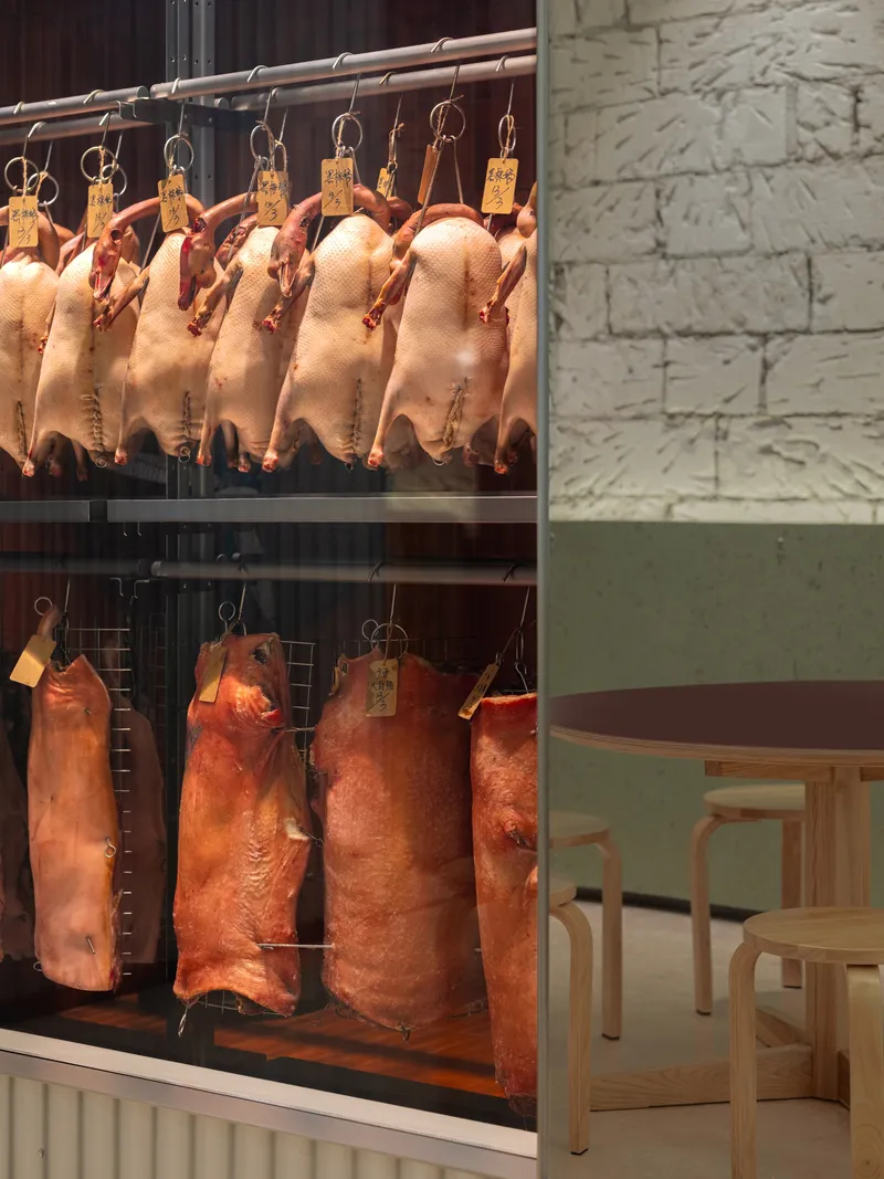 Display window with hanging roasted meats beside seating area with light wood furniture and painted brick wall