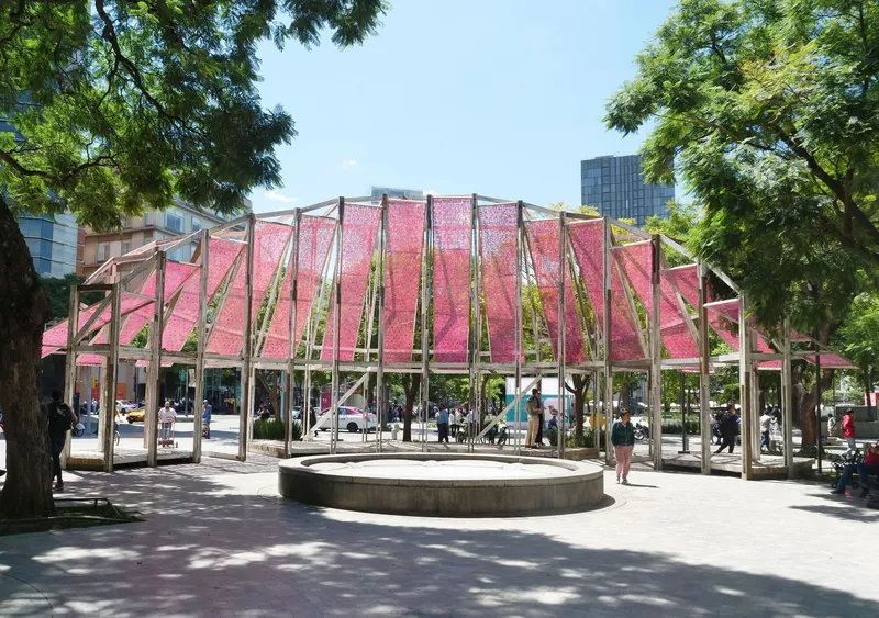 Open timber pavilion with pink fabric roof panels framing a circular concrete fountain under dappled tree shade