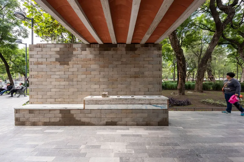 Stone block bench and wall under exposed timber rafters beside a park pathway with a pedestrian passing