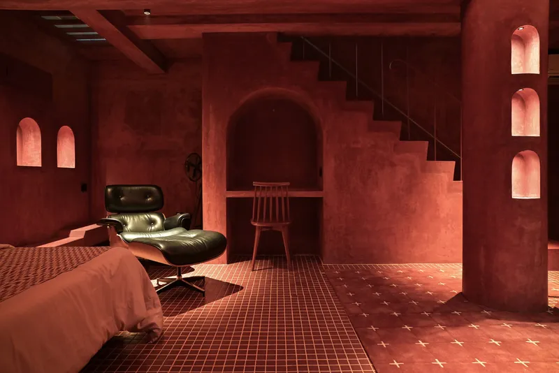 Red plaster interior with arched niches, metal staircase, and cross-pattern tile floor beneath timber ceiling beams