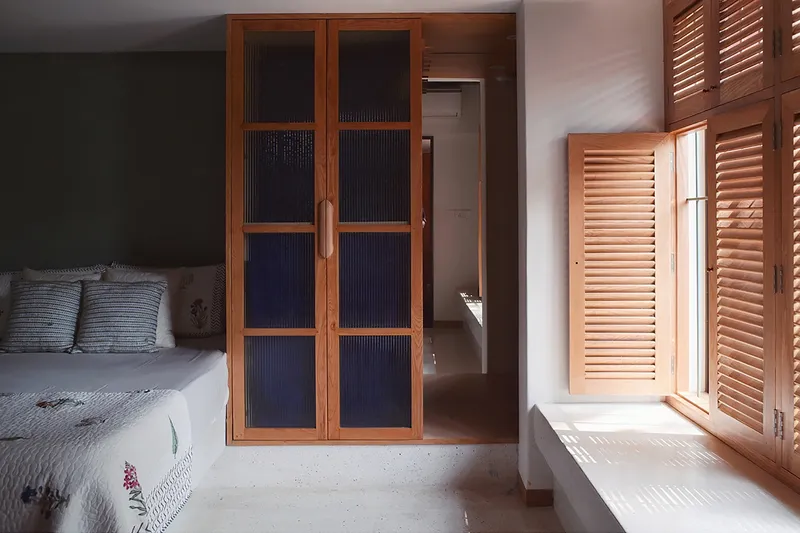 Bedroom with timber-framed glass doors and louvered shutters filtering daylight onto pale concrete floor