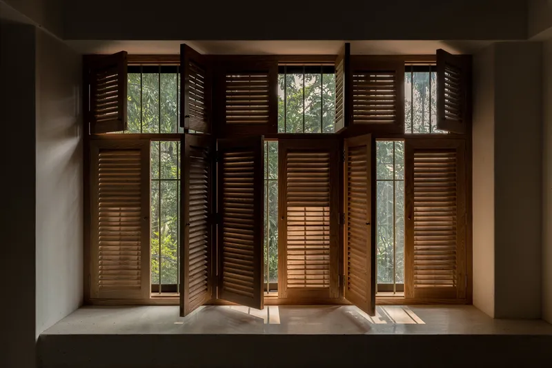 Six timber louvered shutters folded open at window alcove revealing trees beyond in soft daylight