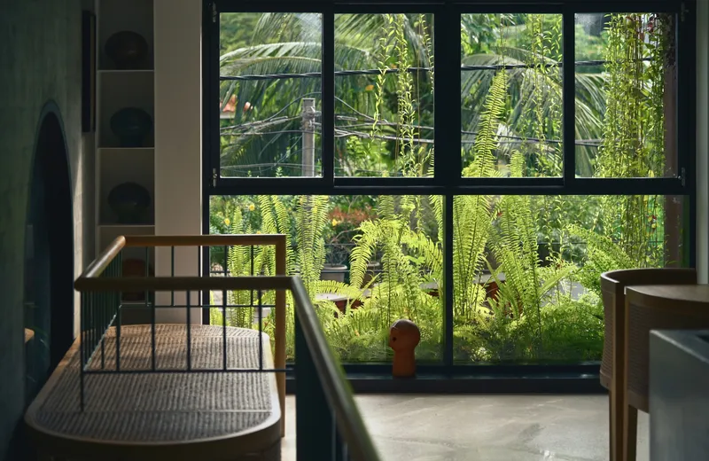 Interior view through black steel-framed windows overlooking dense ferns and tropical vegetation