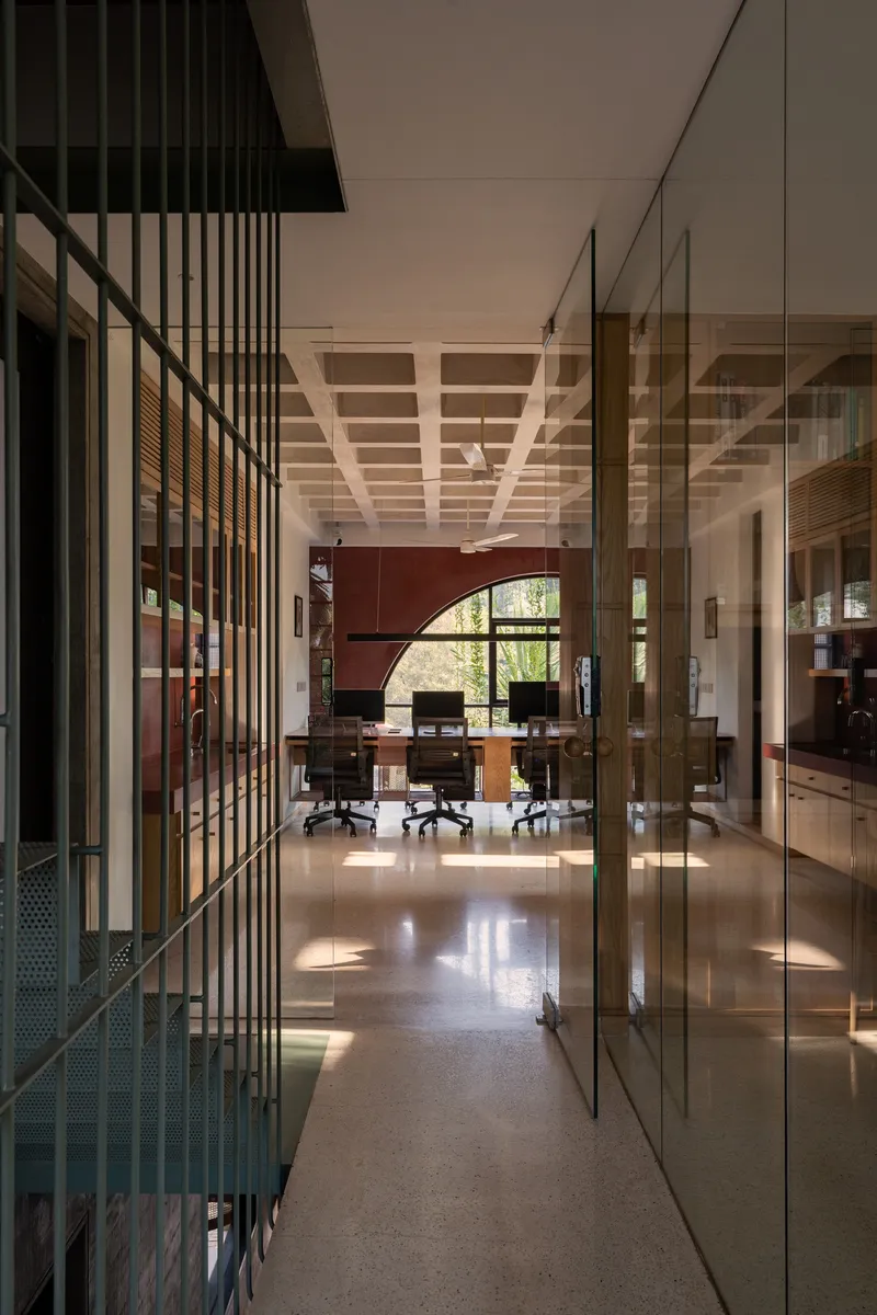 Glass-partitioned corridor with perforated metal screens leading toward arched window and workspace