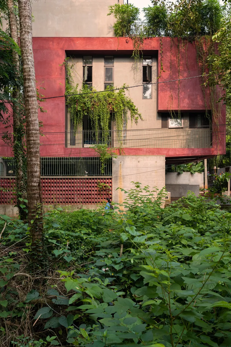 Street view of the layered facade with red oxide panels and concrete base surrounded by dense vegetation