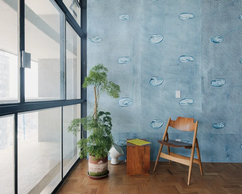 Blue mosaic tile wall with fish motifs beside a steel-framed window and wooden chair with potted plants