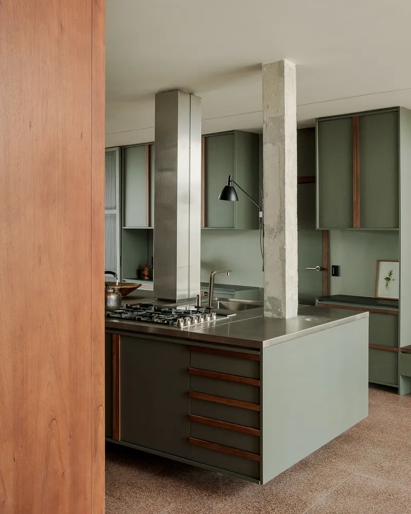 Kitchen island with gas cooktop and exposed concrete column dividing timber and green lacquered cabinetry