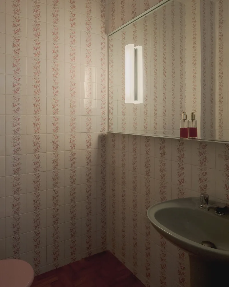 Powder room with floral patterned tile walls and corner mirror illuminated by a vertical wall sconce