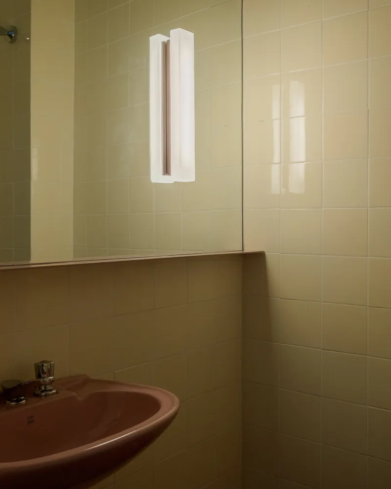 Yellow tiled bathroom corner with mirror and wall-mounted tubular light fixture above a pink sink