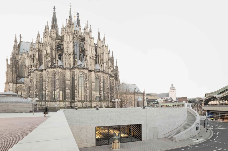 Curved concrete plaza with mesh-screened opening set into the retaining wall below the cathedral