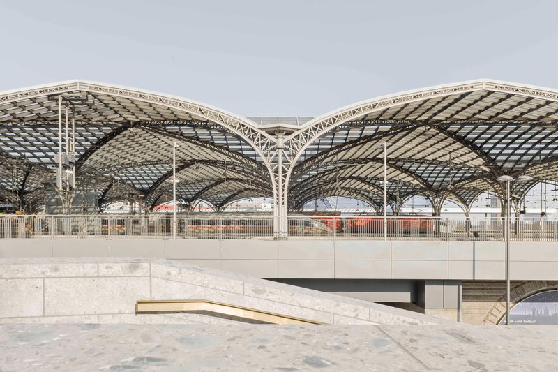 Arched lattice roof structure spanning above a terraced concrete plaza in diffused daylight