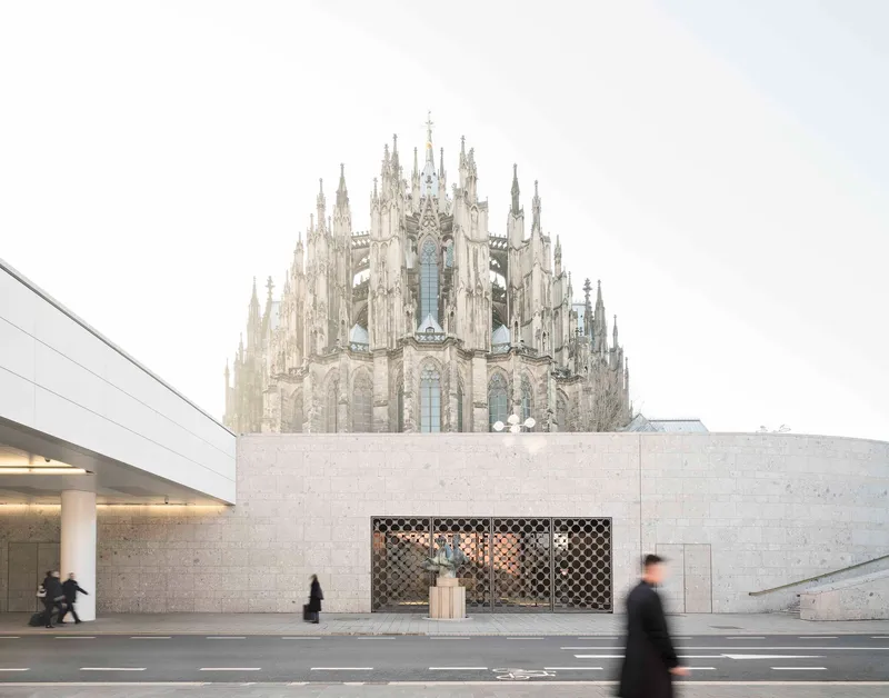 Low stone facade with patterned metal screen framing a courtyard beneath a gothic cathedral on an overcast day