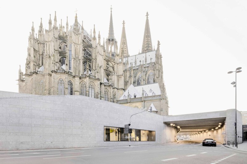 Illuminated tunnel entrance below limestone walls with a gothic cathedral rising behind under grey skies