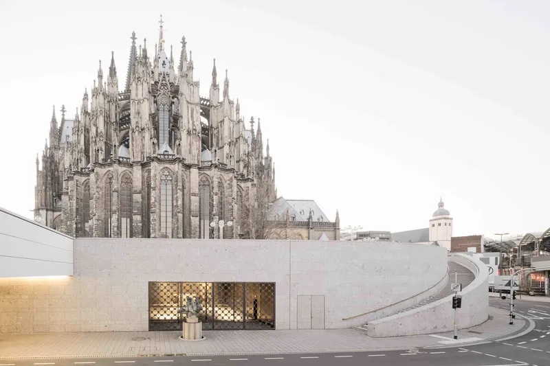 Street view of stone facade with patterned screen windows beneath a gothic cathedral and overcast sky