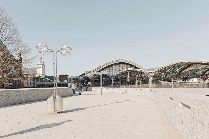 Curved pedestrian plaza with granite pavers leading toward the vaulted station canopies under an overcast sky