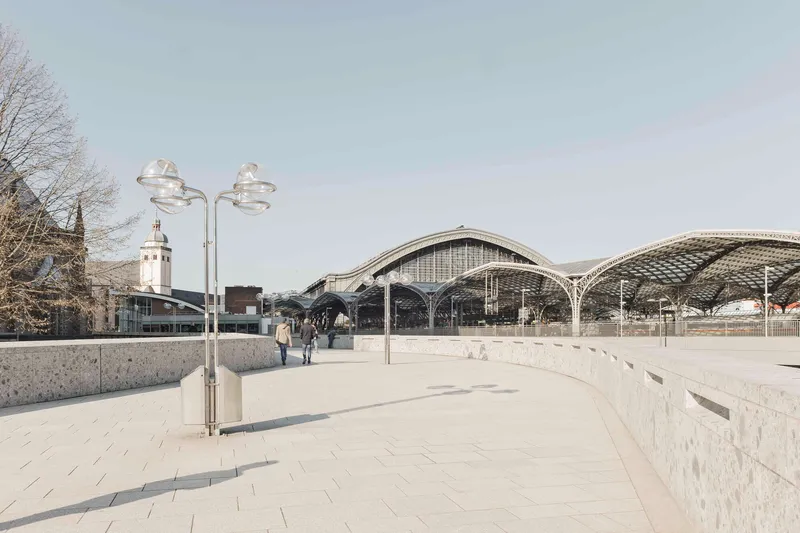 Curved pedestrian plaza with granite pavers leading toward the vaulted station canopies under an overcast sky