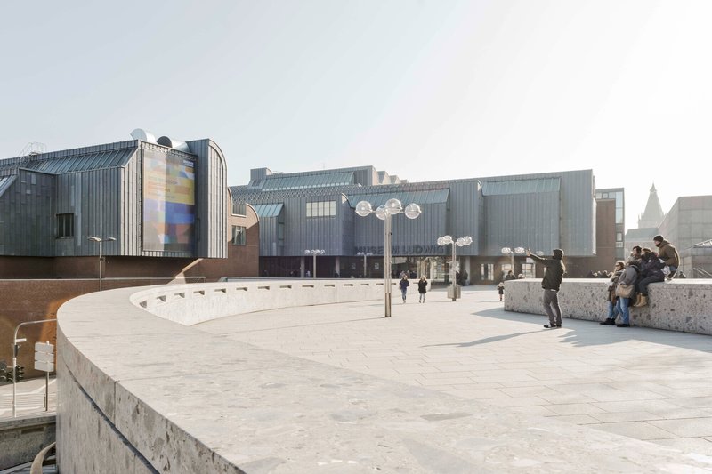 Elevated plaza with concrete benches and paving where visitors gather near the corrugated metal facade
