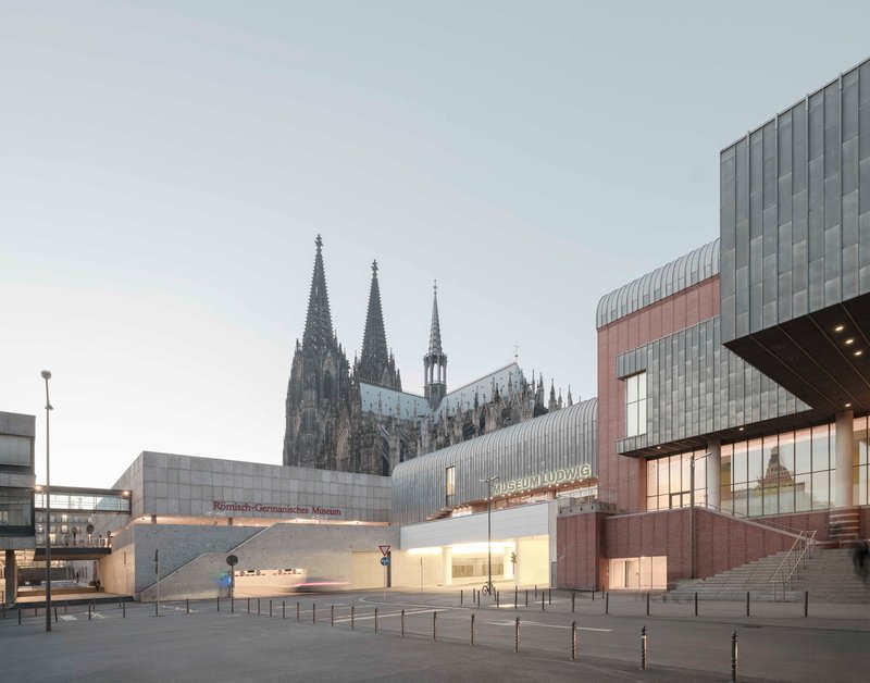 Street view of the ribbed metal entrance volume with illuminated signage at dusk beside the cathedral spires