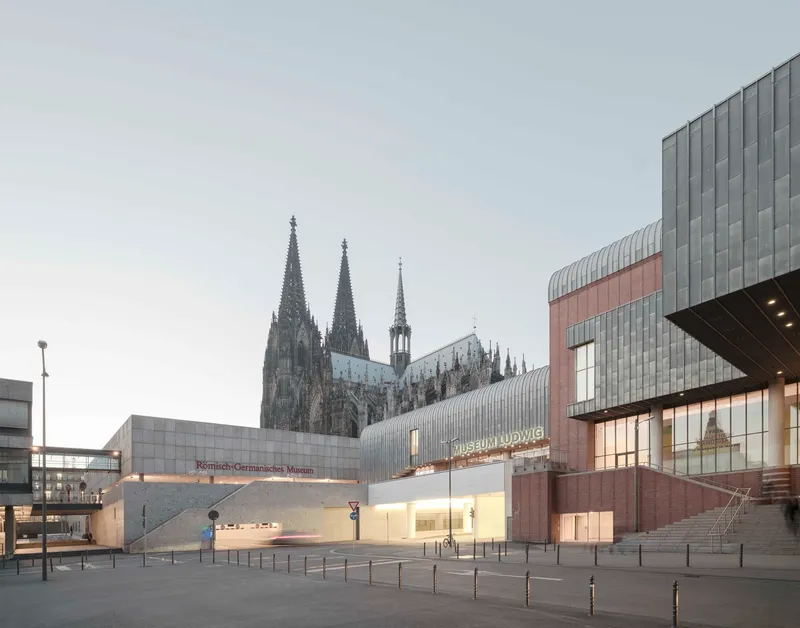 Street view of the ribbed metal entrance volume with illuminated signage at dusk beside the cathedral spires