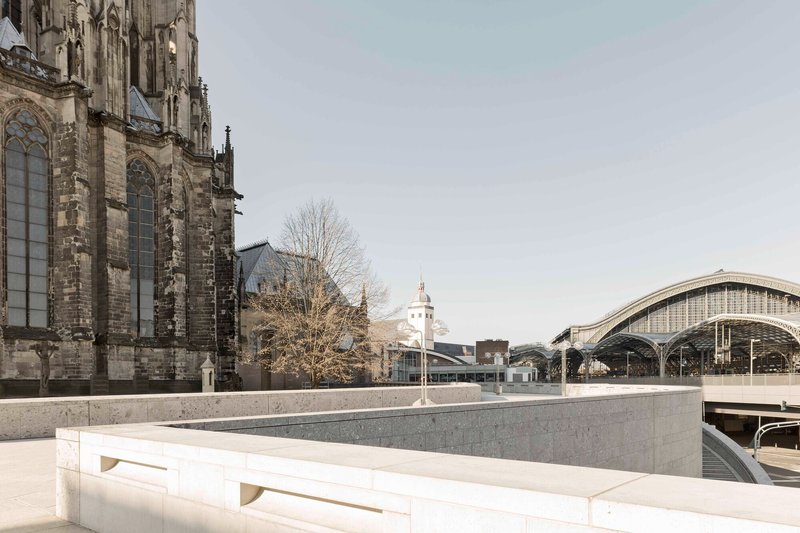 Terraced concrete plaza with linear benches framed by a gothic cathedral and arched train shed