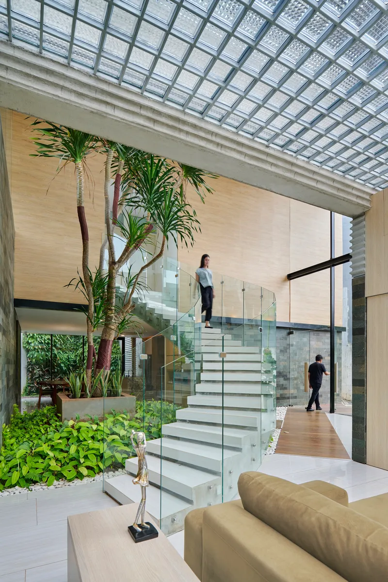 White staircase with glass railings ascending through a double-height atrium with gridded skylight and planted beds