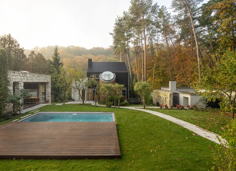 Garden view showing the pool deck, stone pavilion, and black timber volume among tall pines in autumn light