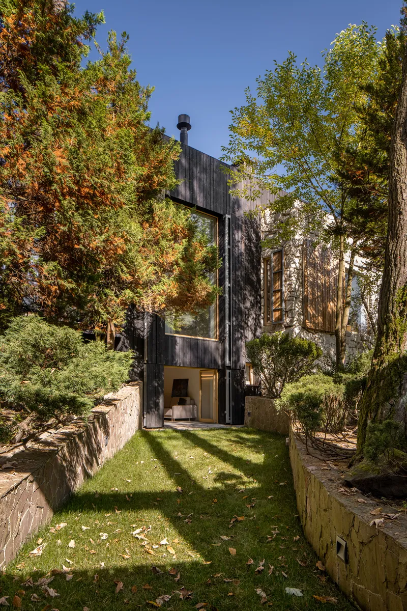 Courtyard view of the dark timber volume rising between stone retaining walls with dappled tree shadows