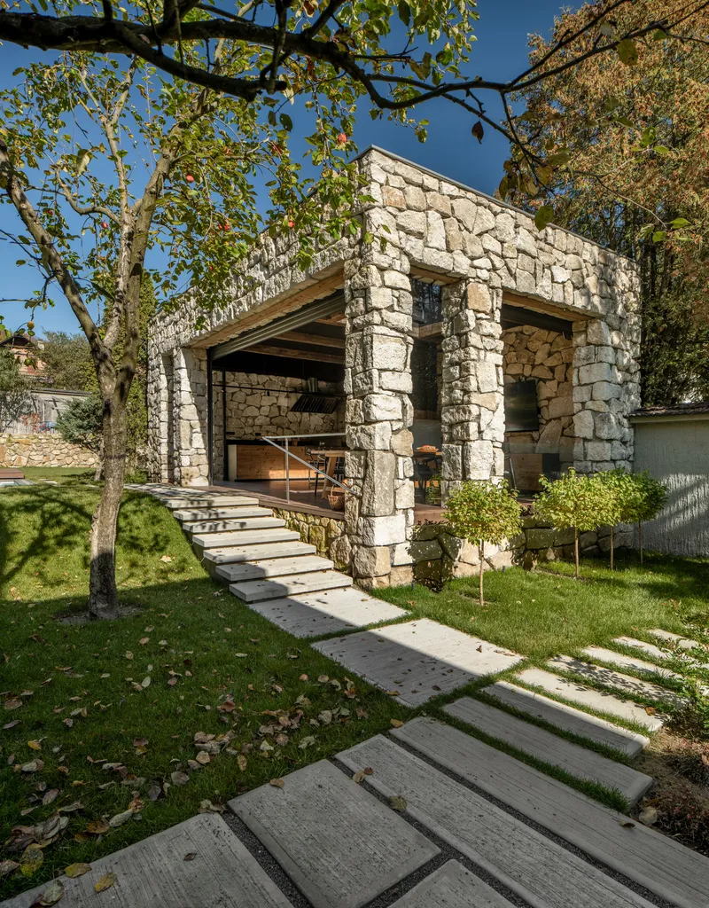 Stone pavilion with exposed timber roof structure and concrete slab path stepping through the lawn