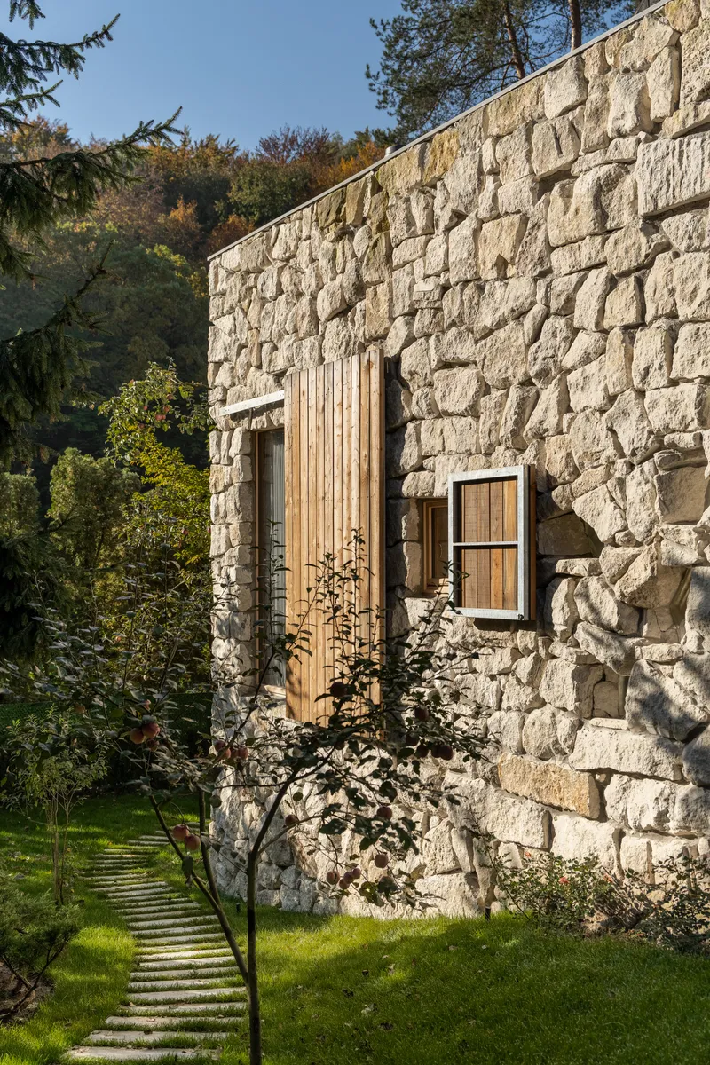 Rough stone wall punctuated by vertical timber cladding and window opening beside stepping stone path