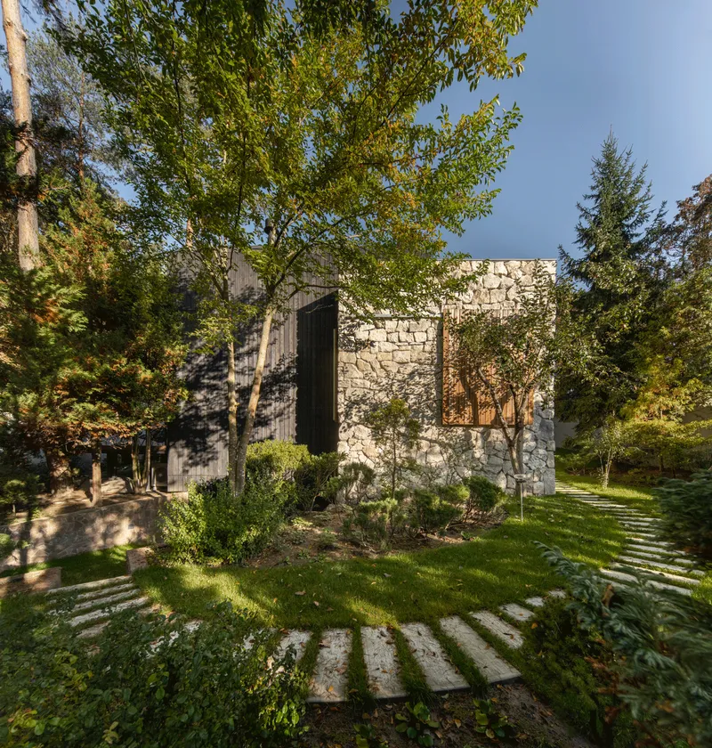 Stone-clad volume with vertical window slot and planted garden beds amid deciduous trees in afternoon light