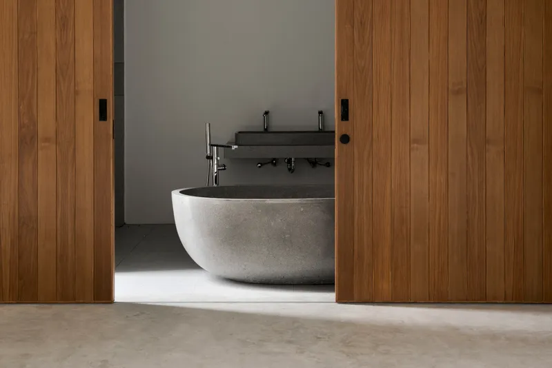 Freestanding stone bathtub framed by timber sliding doors with soft daylight across polished concrete floor