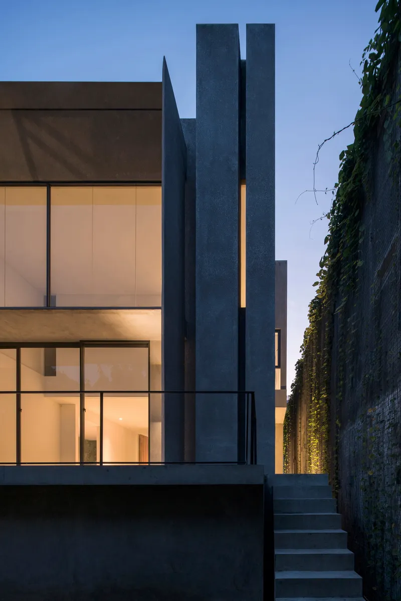 Rear courtyard showing stacked glass volumes beside a tall chimney and illuminated vertical wall planting at dusk