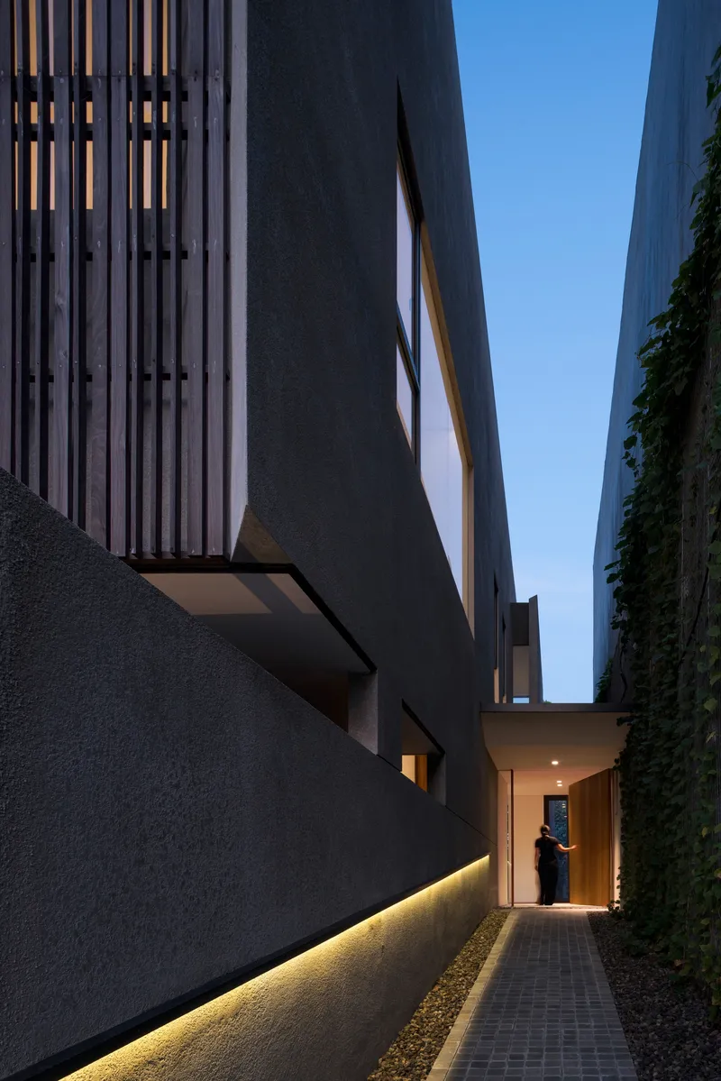Narrow entry passage with ground lighting along the base wall leading to timber door at dusk