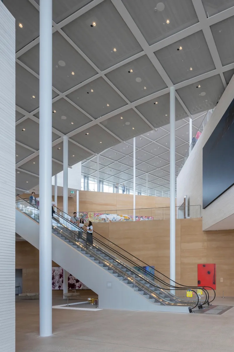 Interior atrium with escalators rising between white columns under a grey coffered ceiling with recessed lighting