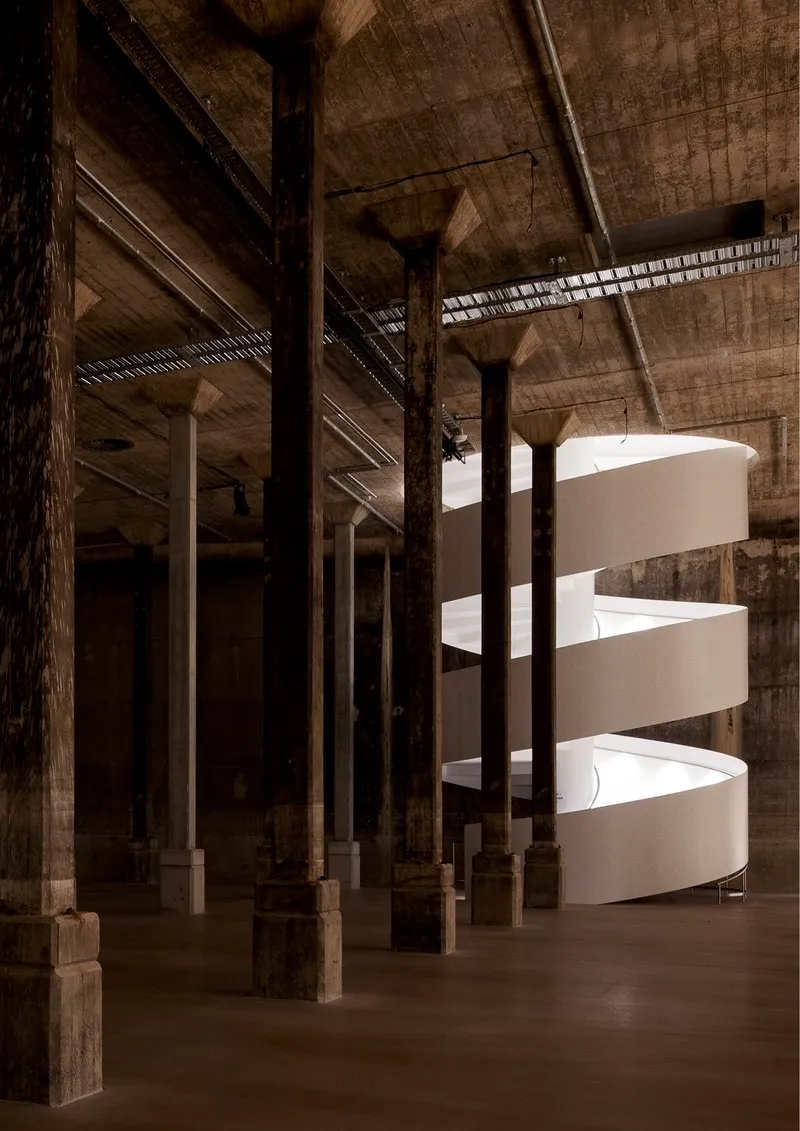 White helical staircase illuminated by clerestory slot within a raw concrete hall with timber columns